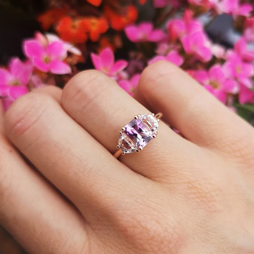 Woman's hand with Lilac Roots ring on the engagement finger, bending the fingers with flowers in the background. Order yours now.