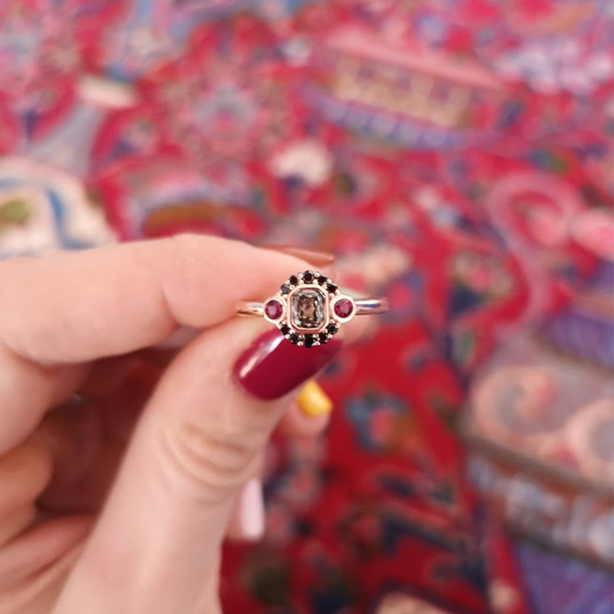 Woman holding La Couche du Diable ring from Lico Jewelry in Montreal, featuring a unique salt and pepper diamond surrounded by black diamonds and rubies set in 18k rose gold, with red nail polish and a beautiful carpet in the background