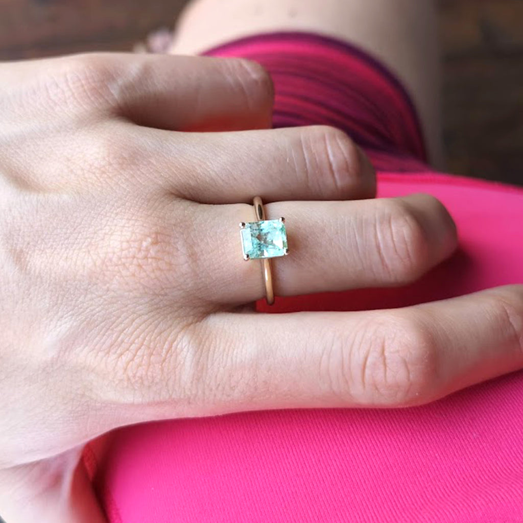 Lico Jewelry's Mint Solitaire ring with 1.57 ct Colombian emerald on woman's middle finger over a pink fabric background.