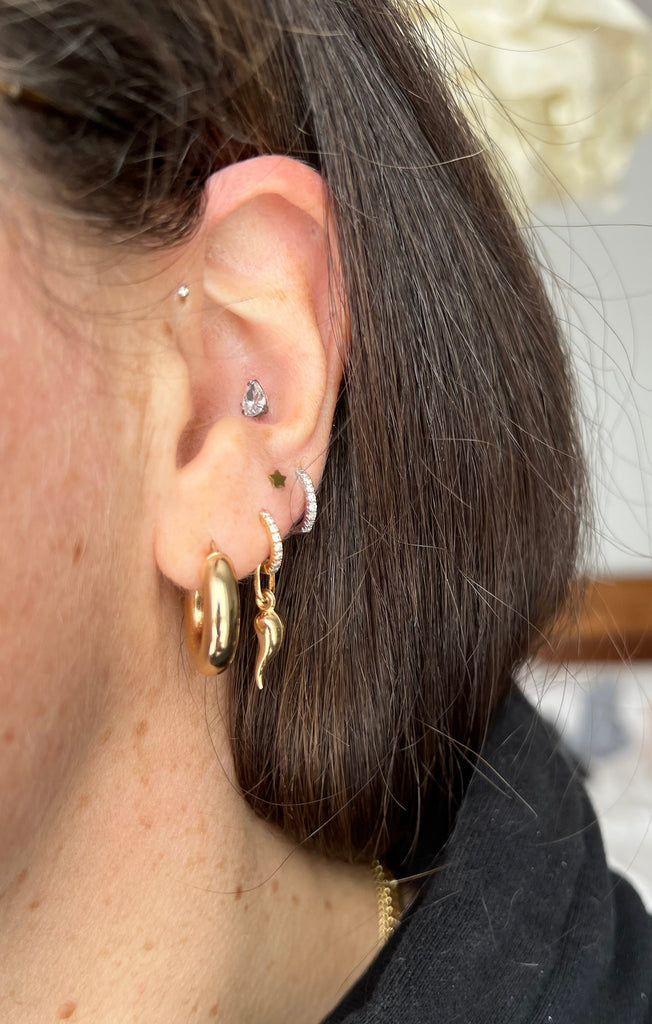 A close-up of a woman wearing a Lico Jewelry's 10k yellow gold Cornicello charm earring as part of a mixed hoop and stud earring set, based out of Montreal.