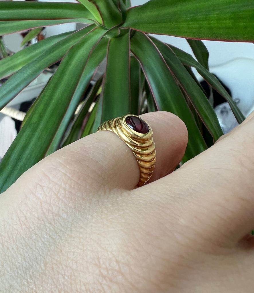Hand model wearing the vintage 18K cabochon garnet ring on the pinky finger, with a lush green plant in the background bending fingers