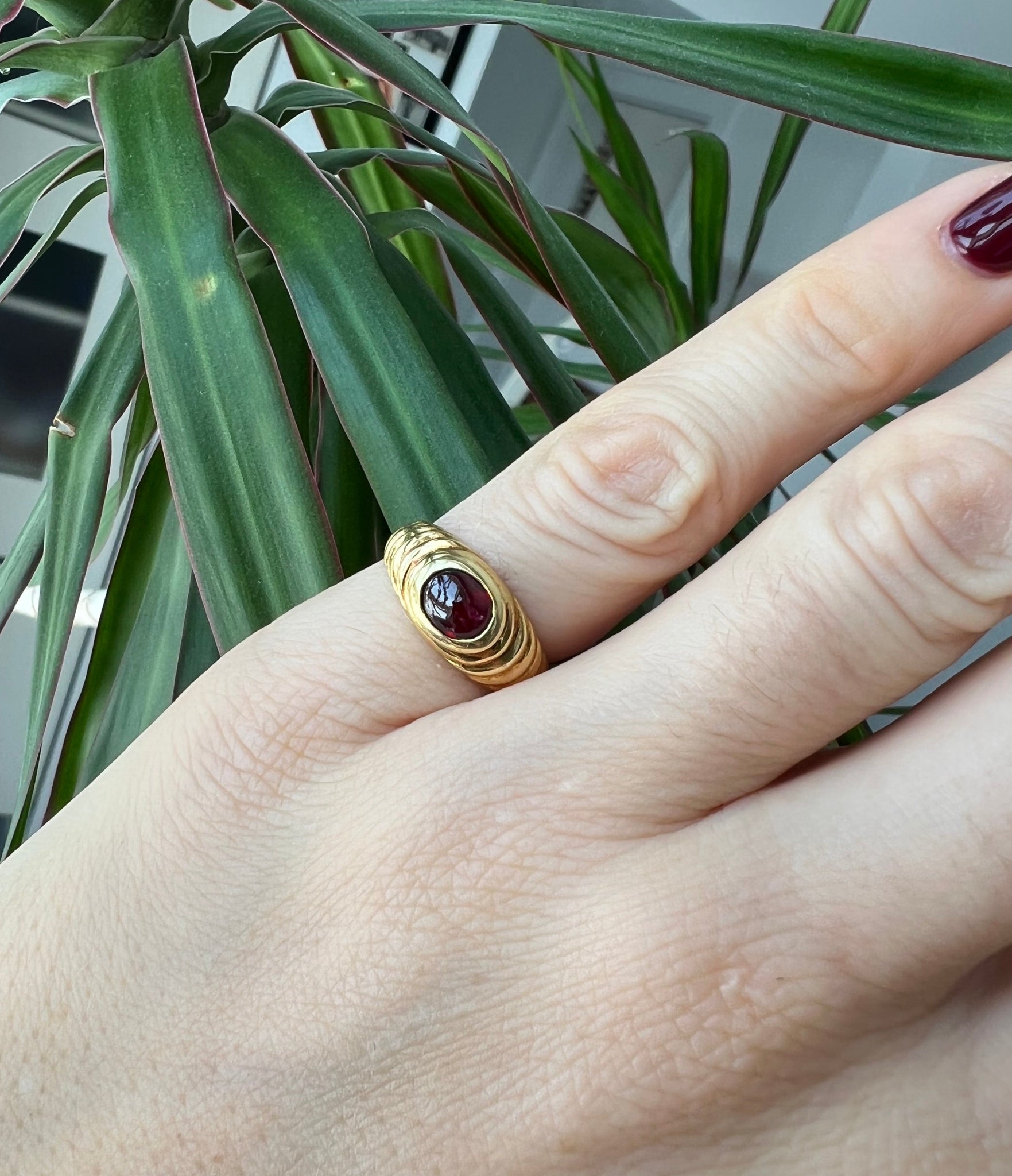 Hand model wearing the vintage 18K cabochon garnet ring on the pinky finger, with a lush green plant in the background.