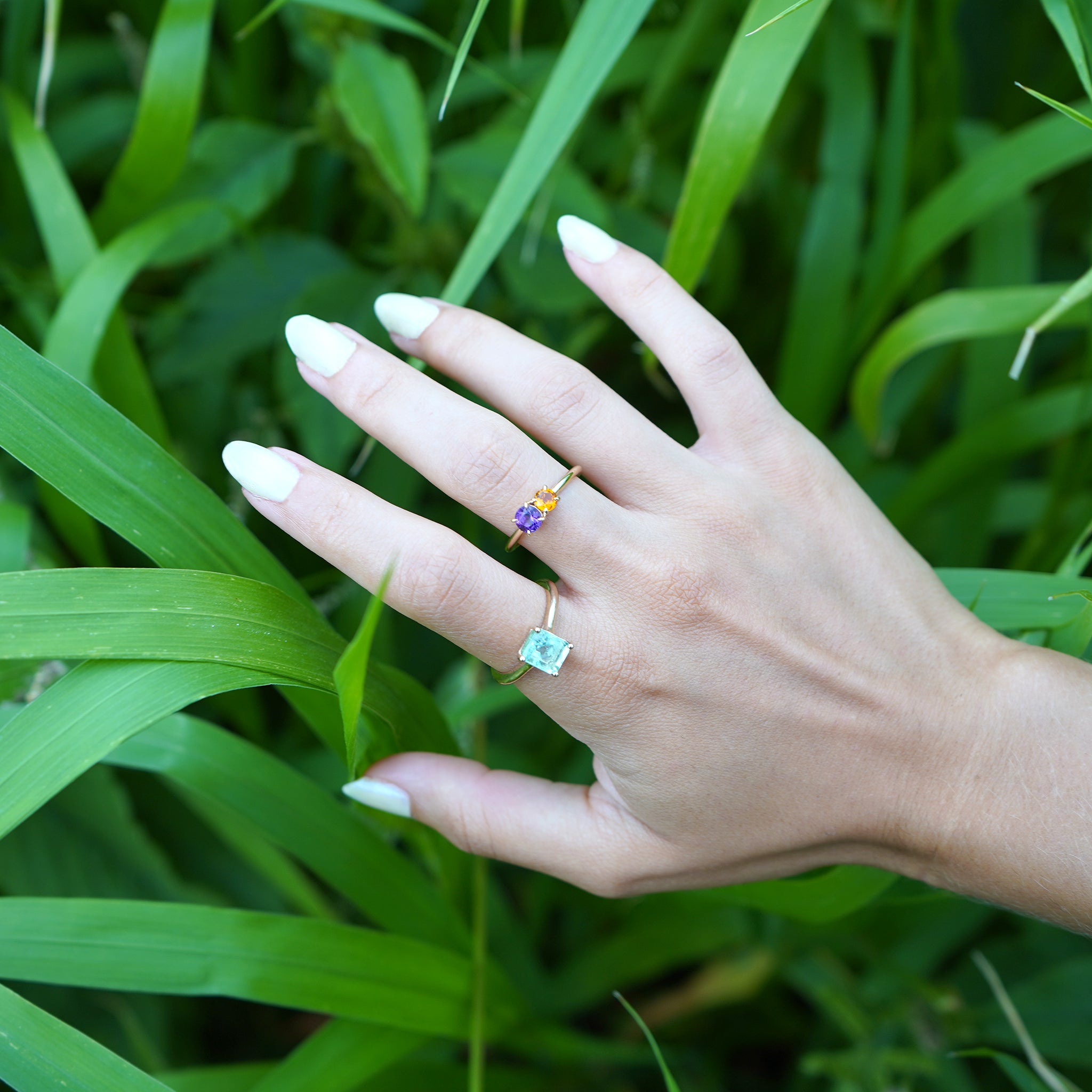 Woman showing off her stylish layered ring look featuring the Iris Flower Ring in solid 14k yellow gold with genuine amethyst and citrine from Lico Jewelry Montreal paired with other rings, set against a background of green plants